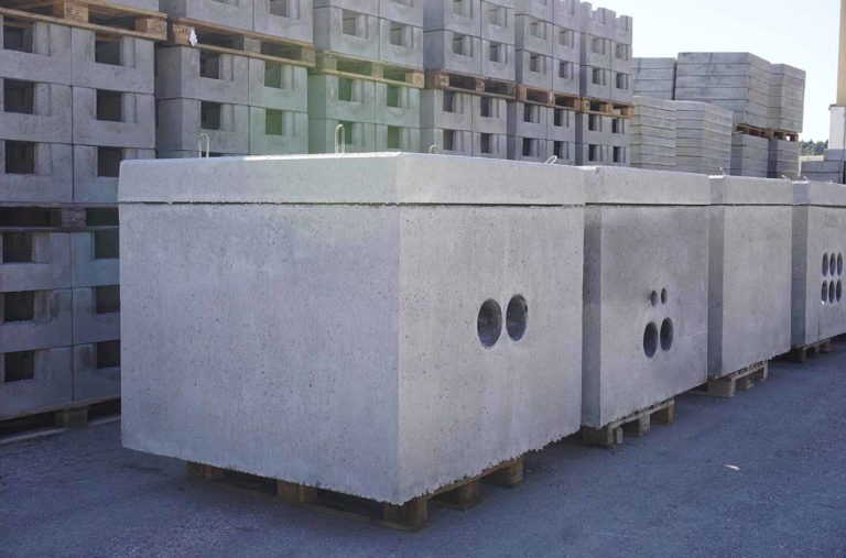 Graue Betonkästen mit runden Öffnungen, gestapelt auf Holzpaletten in einer Lagerumgebung.