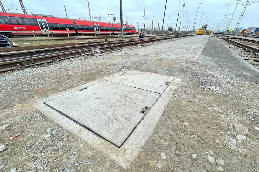 Betonabdeckung für einen Kabelaufbauschacht auf einer Gleisfläche mit Zügen im Hintergrund.