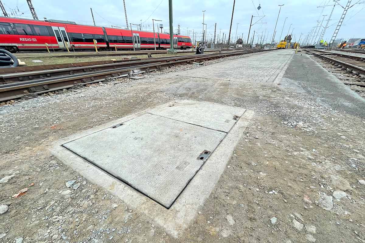 Betonabdeckung für einen Kabelaufbauschacht auf einer Gleisfläche mit Zügen im Hintergrund.