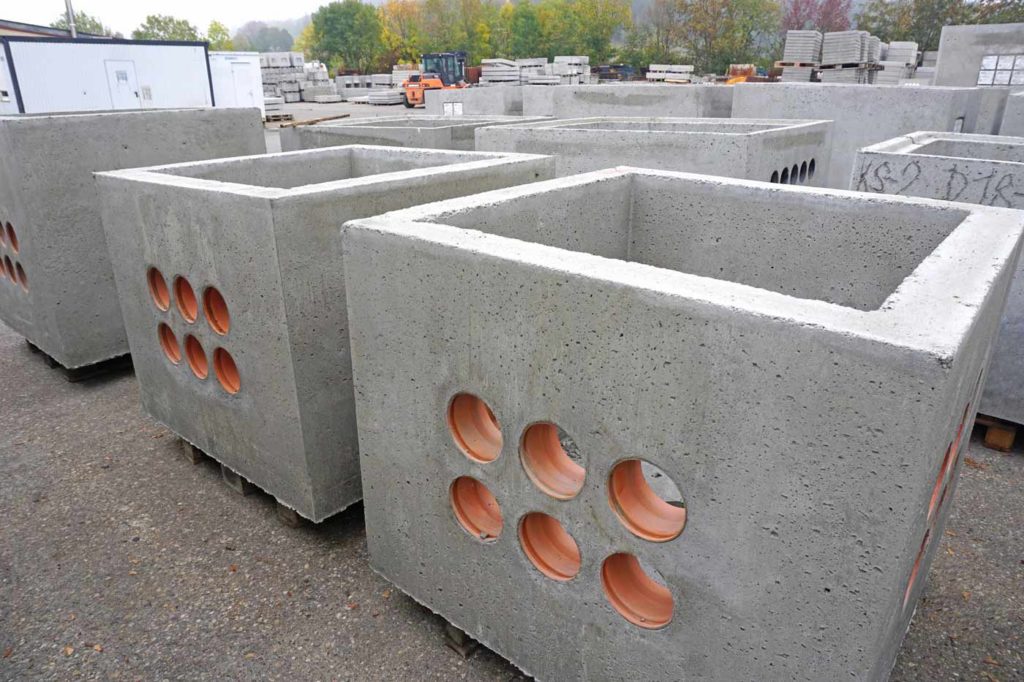 Graue Betonkästen mit runden Öffnungen an den Seiten, in einer Lagerumgebung angeordnet.