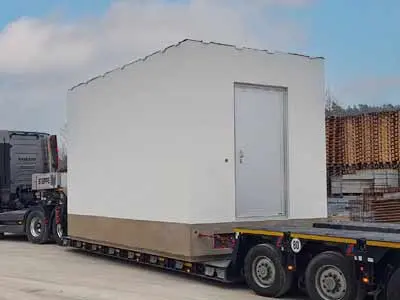 Weißer Baucontainer auf einem Lkw, der auf einer Baustelle transportiert wird.