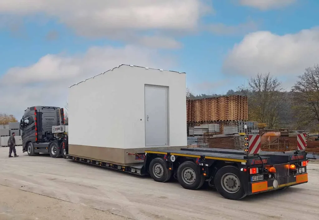 Ein weißer Baucontainer wird auf einem Lastwagen transportiert, mit einer Tür an der Vorderseite.