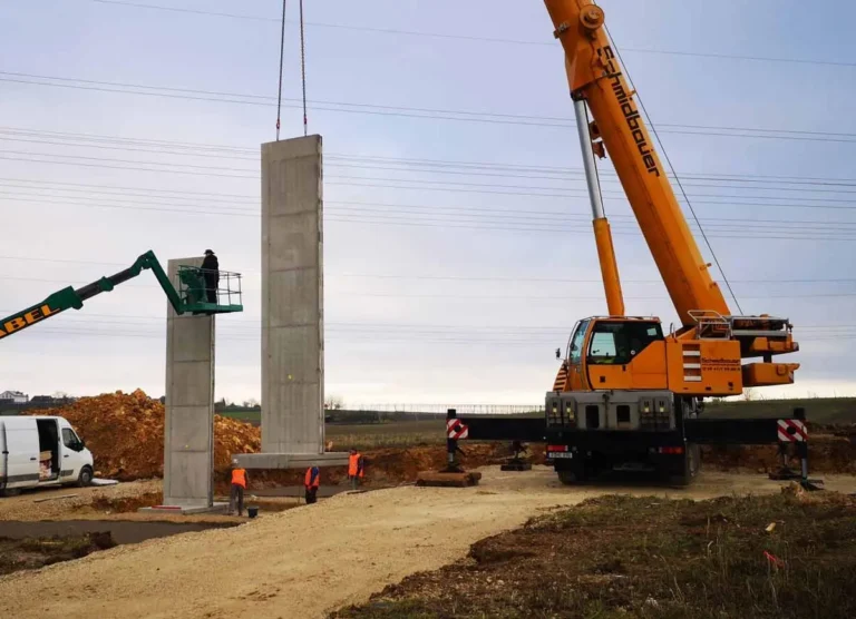 Ein Kran hebt eine Betonwand auf einer Baustelle, während ein Arbeiter auf einer Hebebühne steht.
