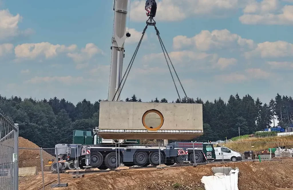 Ein Kran hebt eine große Betonplatte mit einer runden Öffnung auf einer Baustelle.