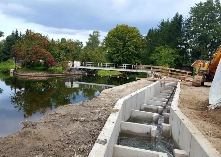 Fischtreppe aus Beton mit Wasserlauf neben einem Gewässer und einer Brücke im Hintergrund.