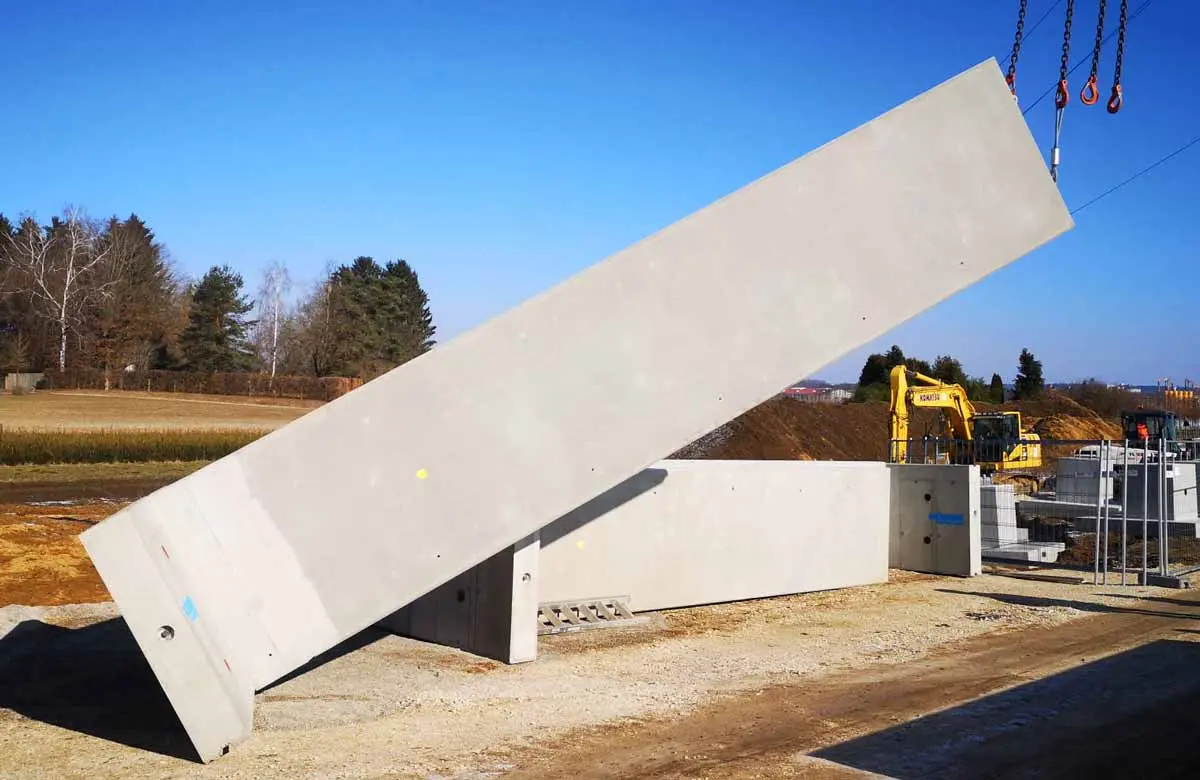 Ein schräg stehendes Betonbauteil auf einer Baustelle mit weiteren Betonplatten im Hintergrund.