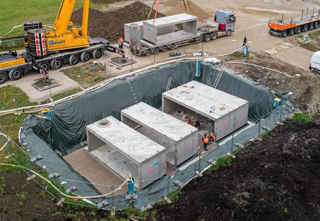 Drei Betonbehälter in einer Baugrube, umgeben von Baumaschinen und Transportfahrzeugen.