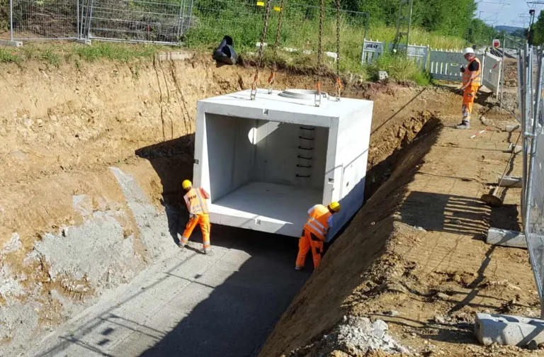 Ein Betonbauwerk wird mit einem Kran in eine Grube gehoben, während Arbeiter in orangefarbener Kleidung assistieren.